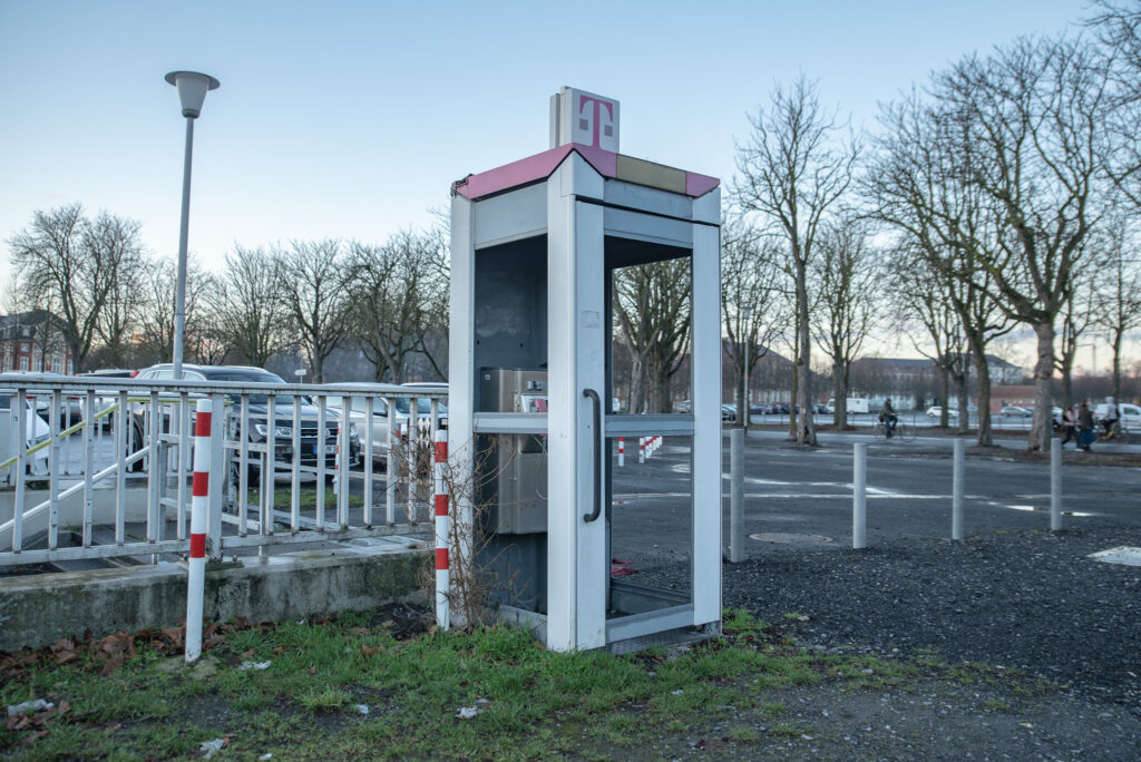 Telefonzelle, im Hintergrund ein Parkplatz und eine Baumallee