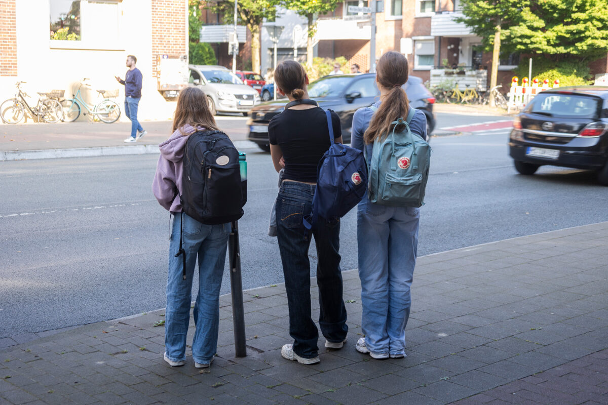 Drei Mädchen stehen mit dem Rücken zum Betrachtenden an einer Bushaltestelle.