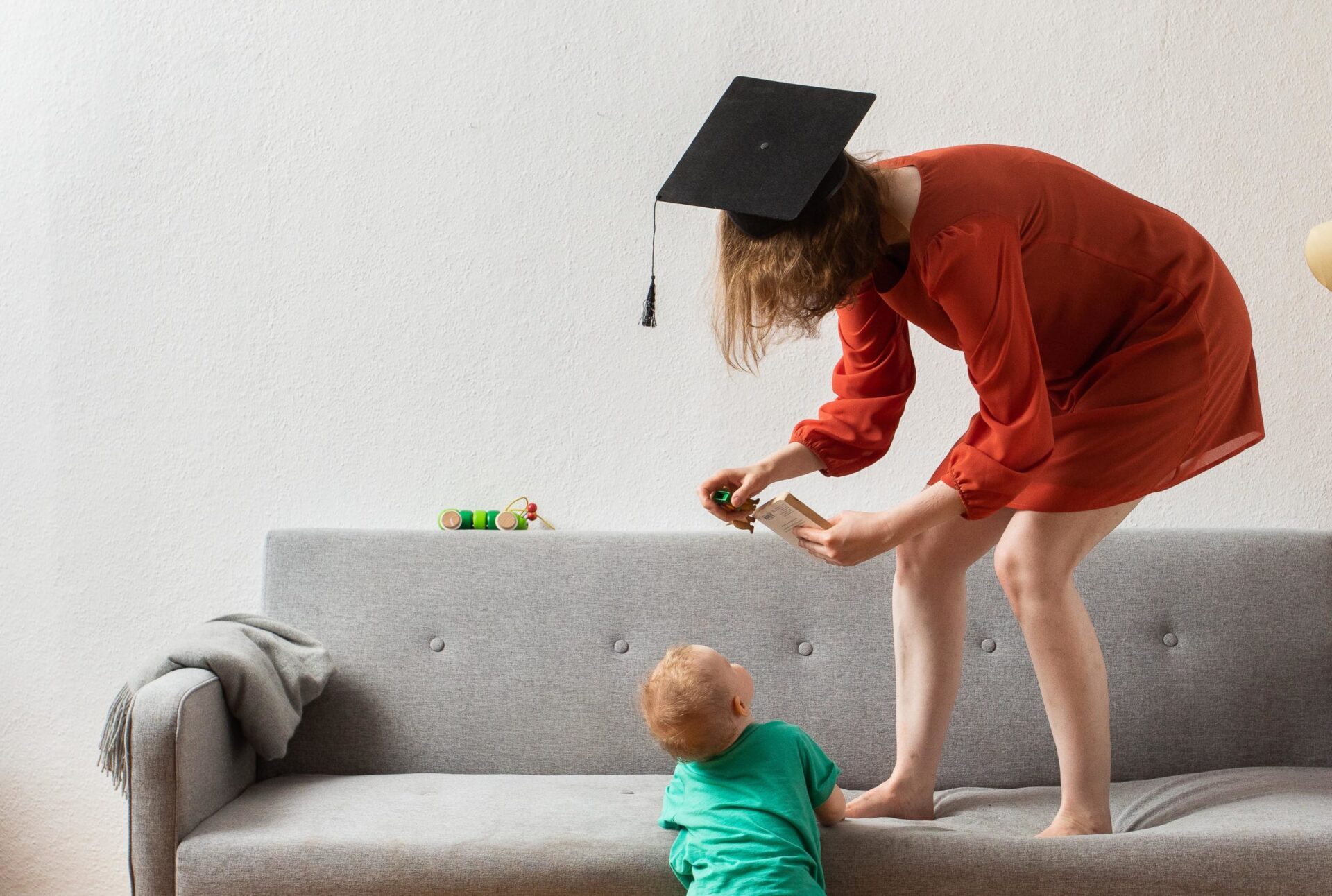 Foto einer Frau mit Absolventenhut, die auf einem Sofa steht und sich zu einem Kleinkind herunterbeugt