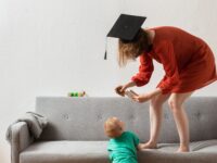 Foto einer Frau mit Absolventenhut, die auf einem Sofa steht und sich zu einem Kleinkind herunterbeugt