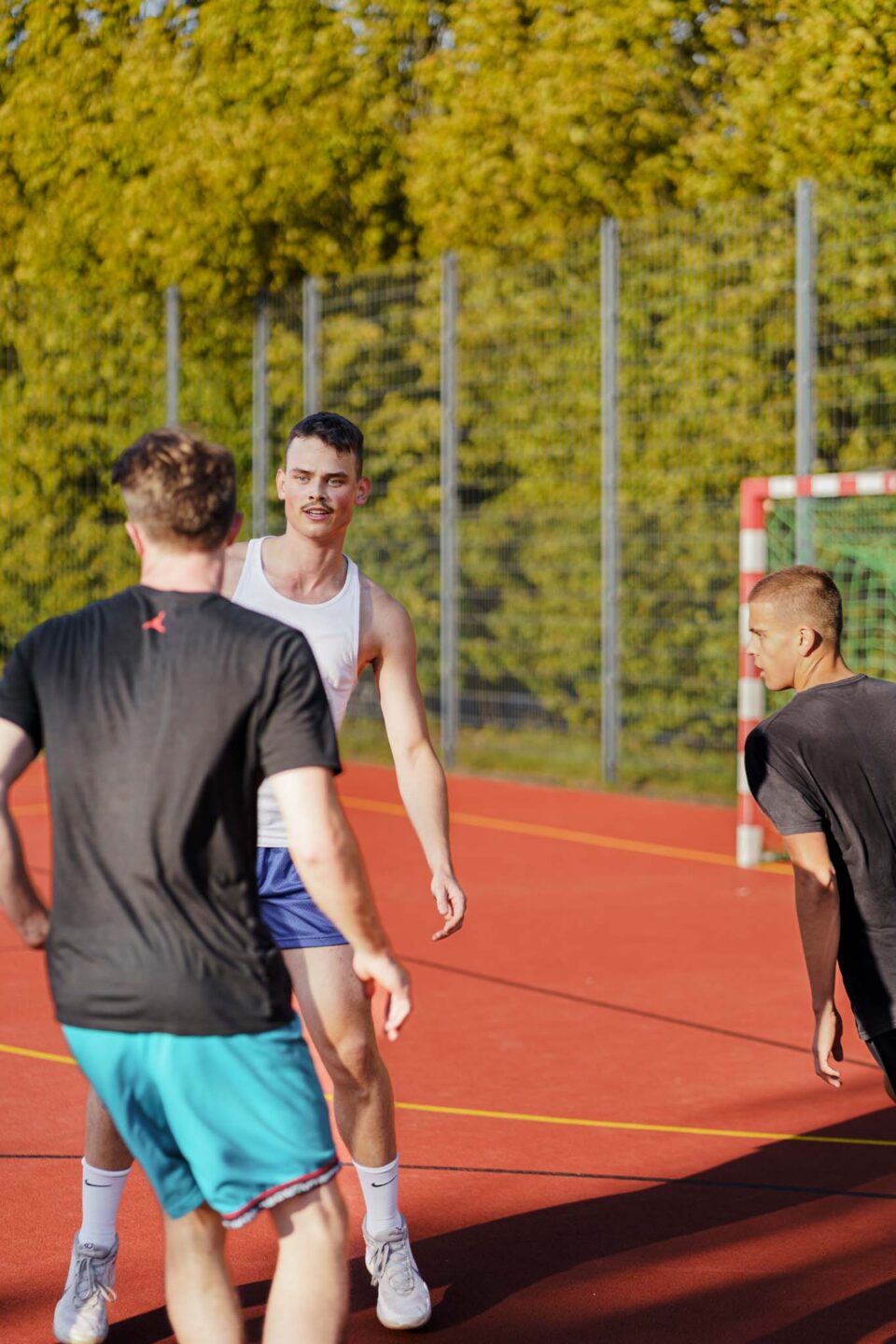 Basketballer in weißem Shirt im Fokus, zwei im schwarzen Shirt im unscharfen Vordergrund. Außenspielfläche.
