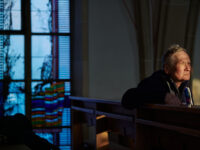 Speck sitzt in einer Sitzreihe in der Kirche und schaut in den Lichteinfall an der Kamera vorbei