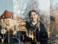 Iris Dzudzek hinter einer Fensterscheibe, in der sich ein Haus spiegelt