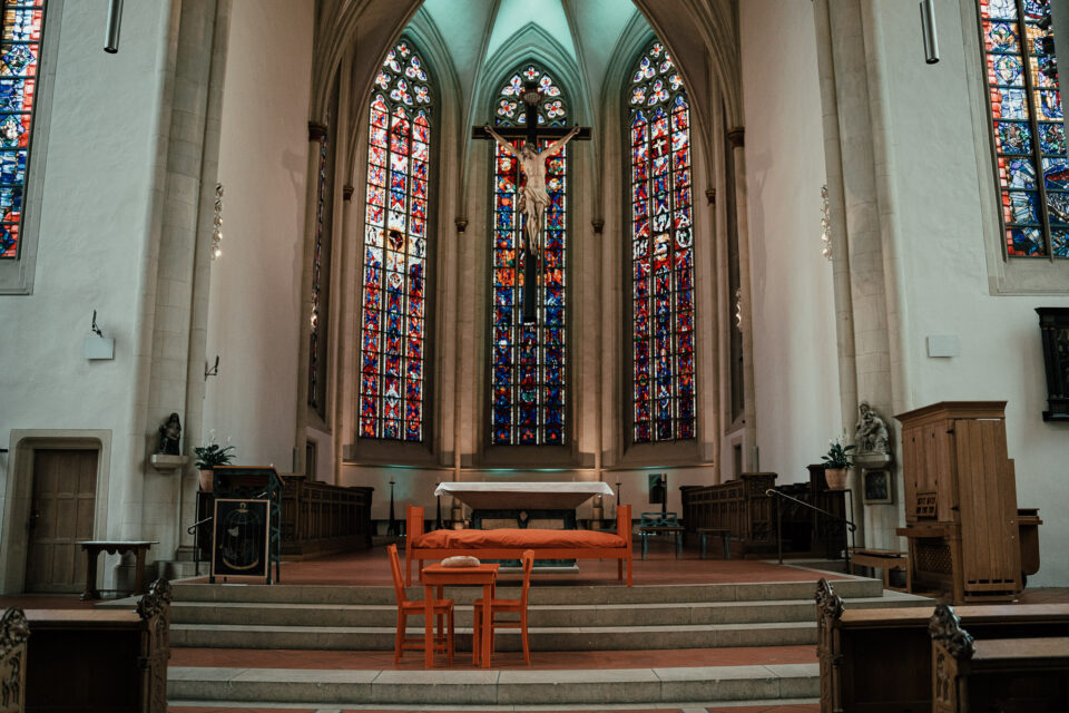 Ein frontaler Blick auf die Ausstellung „Zuflucht geben-gemeinsam hoffen“ in der Überwasserkircher. Es stehen zwei orangene Stühle, ein orangener Tisch und ein orangenes Bett vor dem Altar.